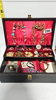 Jewelry box showing open lid with red satin lining holding chains and several bracelets inside compartmentalized sections with various pins and buttons visible