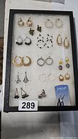 Top view of black framed display case with 16 pairs of assorted earrings on white backing, showing a variety of styles including hoops, dangles, and studs in multiple colors and materials.