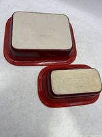 Two Le Creuset rectangular enameled stoneware pans upside down showing beige bottom surfaces stamped with Le Creuset logo and red glazed sides.