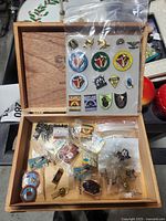 Open cigar box displaying pins organized on a backing board and loose pins in plastic bags inside the box.