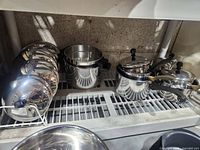 Photo of various shiny stainless steel pots and pans with black handles on a shelf, some stacked with lids.