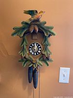 Full front view of the cuckoo clock mounted on a wall, showing the detailed carving of a bird, leaves, and pine cones with clock face and weights.