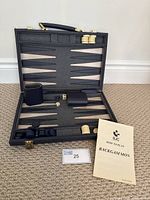 Backgammon game board open in a black carrying case showing playing points, some cream and black playing pieces, dice cups, dice, doubling cube, and an instruction booklet.