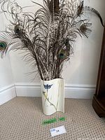Full front view of the Barbara Baatz vase filled with peacock feathers to show size and shape, standing on carpeted floor against white baseboard and wall corner.