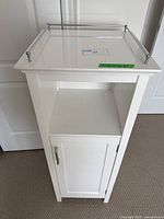 Front view of white cabinet showing top metal guardrails, open middle shelf, and lower cabinet door with metal handle.