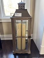 Full front view of the decorative floor lantern showing details of the frame, glass panels, metal roof with ventilation holes, finial top, and large pillar candle inside.