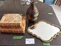 View of two faux book boxes, textured copper-colored vase, and gold-tone scalloped edge mirror on countertop.