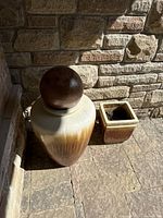 Photo of large urn with attached dark brown spherical ball and small square urn, placed next to stone wall on porch floor.