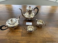 Photo of the silver plated tea set on a wooden table showing teapot, coffee pot, creamer, sugar bowl with black handles.