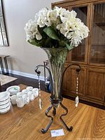 Front view of the glass and metal table vase with white artificial hydrangea flowers inside. The vase is placed on a wood table with a wooden cabinet in the background.