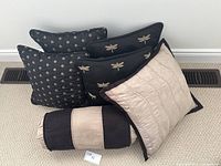 Seven decorative throw pillows displayed on carpeted floor. Four square pillows; two with beige circular patterns on black, two with beige dragonfly embroidery on black; one beige quilted square pillow with black trim; one cylindrical bolster pillow with black and beige stripes.