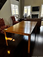Vintage wood dining table shown from an angled side view in a bright room with two red patterned chairs nearby. The table surface shows wood grain and square legs.