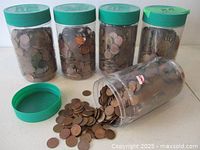 Five clear plastic jars with green lids filled with pennies, one jar tipped showing loose coins.