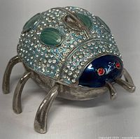 Front and right side angle showing the navy enamel head with red gem eyes and body with clear rhinestones and green oval gems. Silver-tone legs attached.