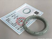 Natural jade D shaped bangle resting on a certification card showing bangle image and authentication details in Chinese