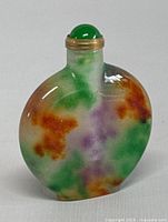 Front view of natural multicolor jade oval snuff or perfume bottle with brass rim and green stopper showing orange, purple, and green accents in the jade stone.