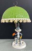 Full view of Bugs Bunny lamp showing gray figurine holding carrot on white base with green textured glass shade with pom-pom trim.