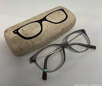 Prescription eyeglasses with translucent gray frames and silver temples next to a beige textured case with black eyeglass image.