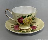 Front view of vintage Queen Anne teacup on matching saucer showing floral pattern with pink and yellow roses, cream background, gold trim and scalloped edges.