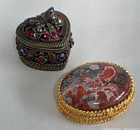 Heart-shaped rhinestone metal jewelry trinket box and oval gold-tone metal pill box with stone top displayed on light surface