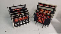 Photo of assorted signs in metal display racks, including Beware of Dog, For Sale, For Rent, Keep Out.