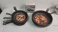 Two stacks of cast iron frying pans on table, labels still intact showing baked items and meat.
