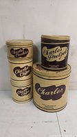Photo showing five vintage Charles Chips tins stacked, including potato chips, pretzels, cookies tins, with cream and brown labels. Condition shows age wear and patina.