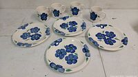 Set of four ceramic plates and four mugs, all hand painted with blue flowers and green leaves on a cream background.