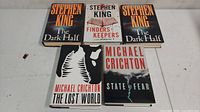 Top view of five novels on a white surface including three by Stephen King (two copies of 'The Dark Half' and one 'Finders Keepers') and two by Michael Crichton ('The Lost World' and 'State of Fear')