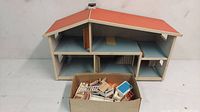 Photo of the vintage wooden doll house with orange roof and box of miniature wooden furniture pieces in front of it