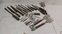 Assorted vintage kitchen knives, forks, spoons, bottle openers, and other utensils laid flat on white surface. Various wooden handles show wear and metal parts have rust.