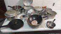 Overview of 11 vintage cookware items on white table including pressure cookers, pots, frying pans, wok, lids, and accessories.