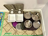 Front view showing the two-bowl timer feeder with white square bowls and dials, along with various metal bowls, flexible lids, and mat arranged on the floor.