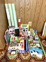 Photo showing assorted crafting supplies laid out on patterned carpet against wood paneled wall. Includes rolls of paper, coloring books, stickers, embellishments, scissors, pins, and foam boards.