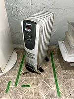 Front angled view of the DeLonghi Magnum electric oil filled radiator heater standing on a tiled floor near a wall and other storage bins. Shows control panel, power cord, and caster wheels.