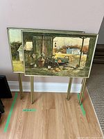 Two folding tables with printed Frank M. Hamilton scenic artwork leaning against a wall showing rustic rural outdoor scenes.