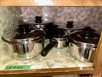 Five stainless steel cooking pots with lids arranged on a shelf inside a cabinet. All pots have black handles and knobs on the lids. The pots appear to be in good condition and vary in size.