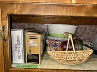 Full shelf view showing boxed Cookie Press and Mixer, plastic bowls, ceramic bowl, and wicker basket and tray