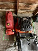 Photo showing Kohler Ariens gas lawnmower with black grass collection bag and two red gas cans placed on a wooden floor in a garage.