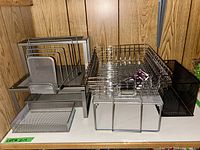 Metal and mesh storage items grouped along wood panel wall. Items include two-tier stand, mail sorter, flat trays, and bins in silver and black metal mesh.