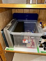 Metal case and clear plastic storage bin containing office supplies on a wooden shelf.