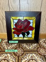 Front view of stained glass rose panel in wooden frame on patterned surface, tags visible with size 13.5 x 13.5 inches and lot number.