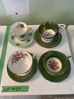 Five teacups and saucers arranged displaying gold and floral detailing and color variations in green and white.