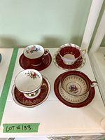 Top view photo showing four red and gold teacups with floral designs inside, each paired with matching saucers on white surface.