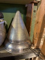 Large large vintage metal cone strainer with handle on shelf with green wall background.
