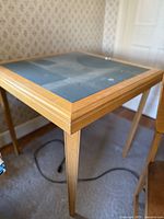 Close-up photo of table corner showing light wood frame with visible wood grain and tapered legs, dark tabletop surface with minor wear.