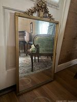 Full view of rectangular mirror with gilded frame and floral basket motif at top, reflecting interior room objects.