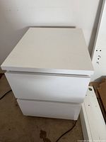 Two white IKEA nightstands each with two drawers, shown from different angles on carpeted floor against white wall.