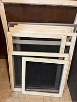 Grouped vintage wood frame window screens stacked on floor against wall, all with mesh and varying sizes.
