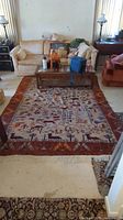 Full view of the rectangular area rug with rust-colored border and animal, people, and nature motifs in the center.